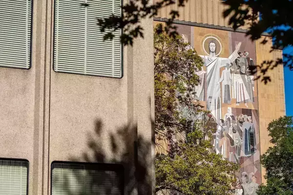 The Word of Life mural on the Hesburgh Library features a mosaic of Jesus with arms raised, surrounded by numerous saints and scholars. Green tree branches partially frame the colorful mosaic and tan building under a blue sky.